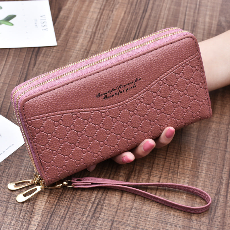 Pink textured wallet with embossed pattern, gold zipper, and wrist strap on a wooden surface.
