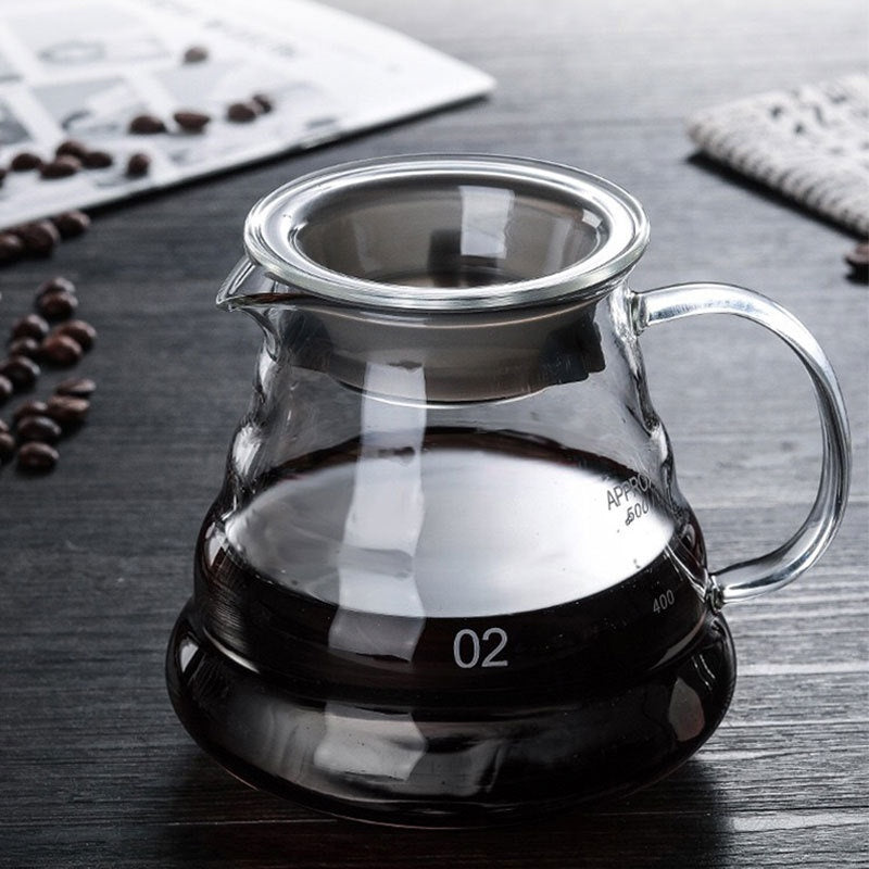 Glass coffee carafe with a handle, filled with dark liquid, on a wooden table beside coffee beans.