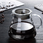 Glass coffee carafe with a handle, filled with dark liquid, on a wooden table beside coffee beans.